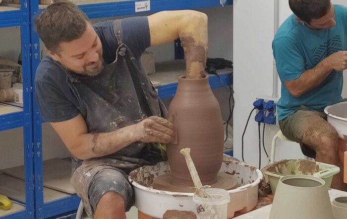 Un hombre da forma a un jarrón de arcilla en un taller de cerámica con torno, rodeados de herramientas para su elaboración.