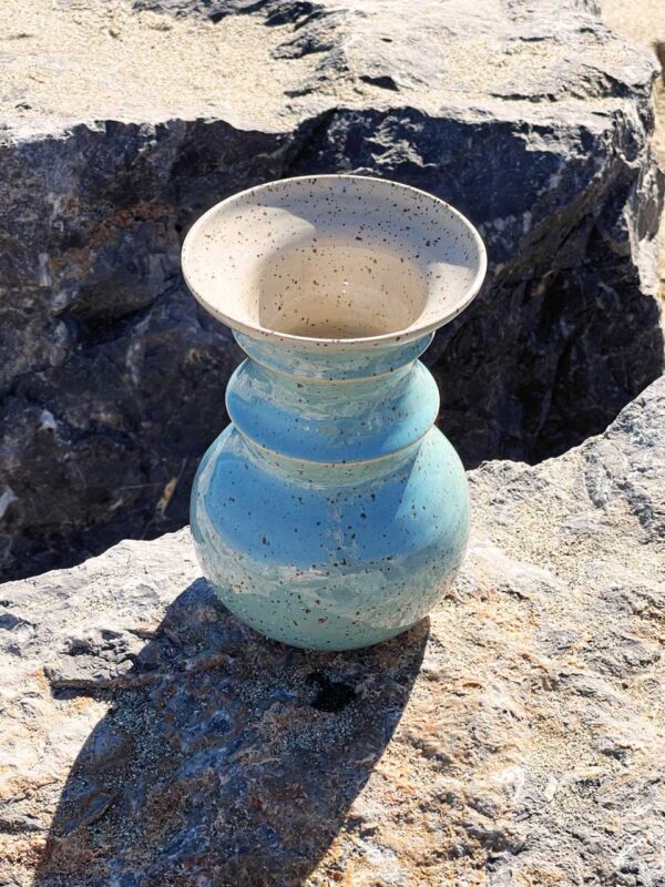 Un florero de barro decorado sobre una roca, con el océano al fondo y la luz del sol iluminando la escena.